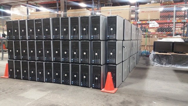 PC Recycling solutions for small businesses. A warehouse scene showing a large stack of black and silver desktop computer towers organized in a neat, grid-like pattern. Orange traffic cones are placed in front of the stack, and various other items are visible in the background, including shelving and equipment